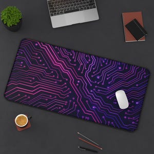 May include: A rectangular mouse mat featuring a circuit board design in purple and pink hues. A white computer mouse sits on the mat. Other items on the dark surface include a laptop, a small potted plant, a notebook, and a cup of coffee.