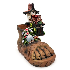 Puede incluir: Una maceta de cerámica marrón con forma de pie gigante, adornada con una casa en miniatura y un jardín. La casa tiene un tejado de tejas rojas y el jardín presenta flores coloridas y vegetación.