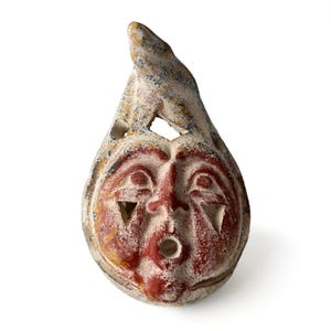 May include: A teardrop-shaped ceramic whistle featuring a hand-painted face with red and white pigments. The object has a textured, weathered surface, triangular cutouts for eyes, and a circular opening for the mouth, with a pointed top section.