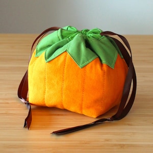 Op de afbeelding: Een kleine oranje pompoenvormige trekkoordtas met een groene bovenkant en bruine lintjes.
