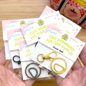 May include: Several packages of tiny hardware for duffles are displayed. The packages have a pink and white design with the text "TINY HARDWARES FOR TINY DUFFLES". The hardware includes gold and black metal clasps and rings.