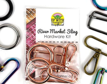 May include: A clear packet labelled "River Market Sling Hardware Kit" contains rose gold metal rings and clasps. Various metal hardware pieces in black, silver, and iridescent colours surround the packet. The brand name "Up With Handmade BanjoFlannel" is visible.