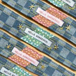 Pode incluir: Close-up de várias tiras de tecido com um padrão xadrez azul. Cada tira tem uma seção de bolinhas rosa e uma seção de bolinhas verde-água. O texto "HELLO, GORGEOUS!" está impresso em uma etiqueta branca. Bordados dourados em forma de estrela acentuam o design.
