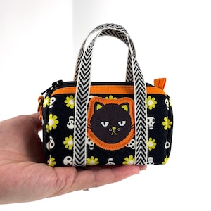 May include: A small, black fabric handbag with a Halloween theme. The bag features a black cat applique, yellow flowers, and skull patterns. It has a black zipper, orange trim, and a chevron patterned handle.
