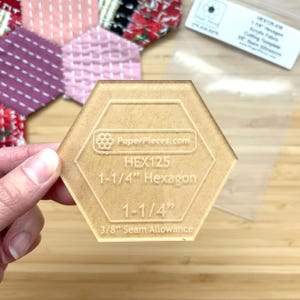 Op de afbeelding: Een doorzichtige, zeshoekige acrylsnijmal met de tekst "PaperPieces.com", "HEX125", "1-1/4" Hexagon", "1-1/4" en "3/8" Seam Allowance". De mal is 3,18 cm breed.