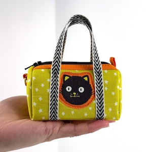 May include: A small, yellow fabric bag featuring a black cat face applique. The bag has a black zip, orange trim, and a white star pattern. The handles are black and white chevron. A charming and whimsical accessory.