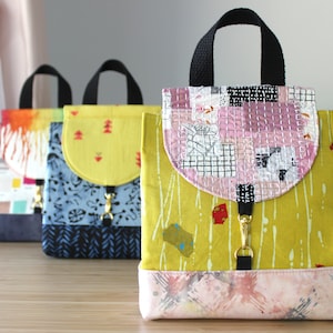 May include: Three colorful fabric backpacks with different patterns. The backpacks have black straps and a flap closure with a gold clasp. The backpacks are on a wooden surface.