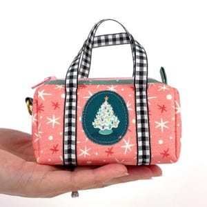 May include: A small, pink fabric duffel bag with a Christmas tree design. The bag features a black and white checkered handle and straps, a teal zipper, and a circular patch with a decorated tree. White snowflakes are scattered across the pink background.