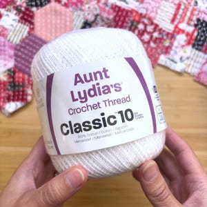 Puede incluir: Un ovillo de hilo de ganchillo blanco Aunt Lydia's Classic 10 con una etiqueta de papel blanco con texto morado. La etiqueta muestra el nombre de la marca, el tipo de producto, el tamaño 10 y especifica que es 100% algodón mercerizado.