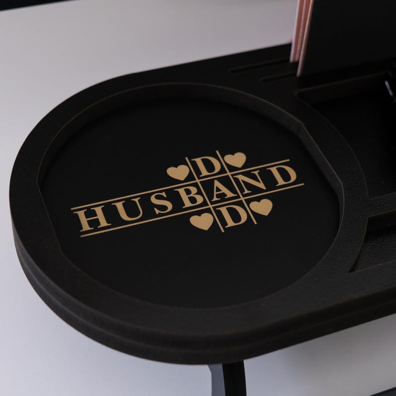 May include: Black desk organizer with a circular tray featuring the gold text "HUSBAND" with heart symbols. The organizer has multiple compartments and a raised edge. The background is white.