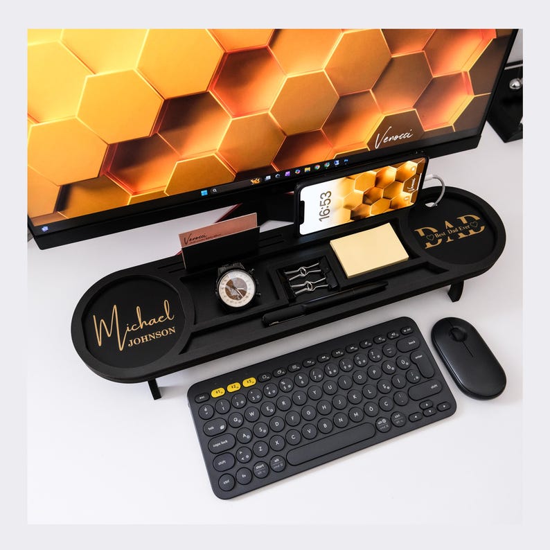 May include: Black desk organizer with a personalized name and the word "DAD" on the sides. It holds a phone, watch, pens, and business cards. A black keyboard and mouse are in the foreground, with a computer monitor in the background.