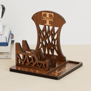May include: A wooden phone stand with a dark brown finish. The stand features a decorative geometric design and the name "Thomas" with an initial "T" engraved at the top. The base has the phrase "I love you?" engraved.
