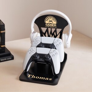 May include: Black gaming controller stand with the text "Thomas" in gold. The stand holds two white and black controllers and a pair of white headphones. The stand has the text "DAD BY DAY GAMER BY NIGHT".