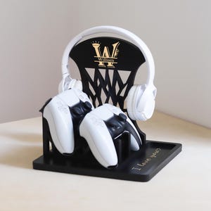 May include: A black gaming headset and controller stand with a gold monogram and the name "William". The stand holds a white headset and two white and black controllers. The base has the text "I love you?"
