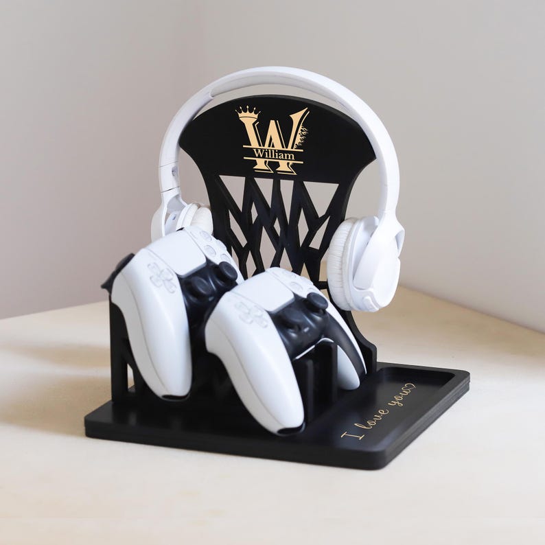 May include: A black gaming headset and controller stand with a gold monogram and the name "William". The stand holds a white headset and two white and black controllers. The base has the text "I love you?"