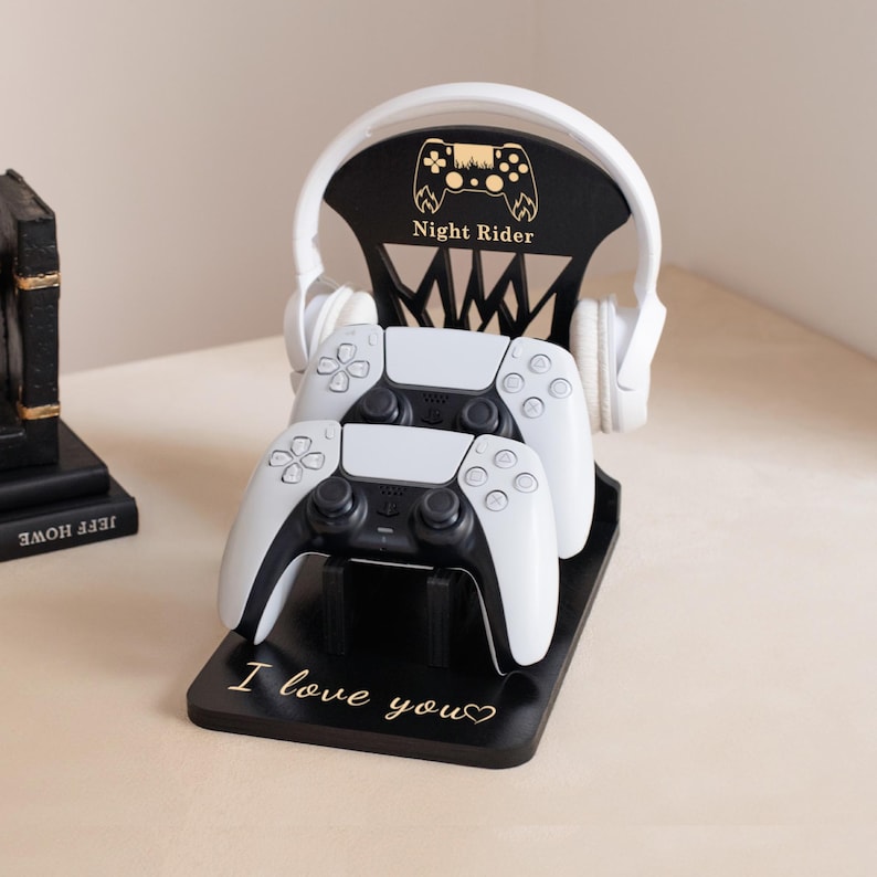 May include: Black wooden gaming controller and headset stand with the text "Night Rider" and "I love you". The stand holds two white and black controllers and a white headset. The stand is on a light-colored surface.