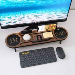 May include: A wooden desk organizer with a phone holder, pen, and notepad. The organizer has two circular areas with initials and the word "DAD". A wireless keyboard and mouse are in the foreground.