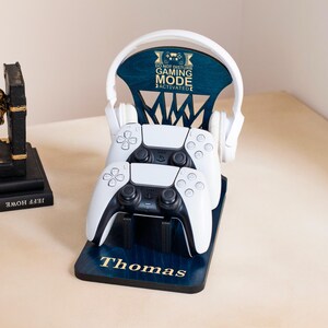 May include: A wooden gaming headset and controller stand in blue with the text "DO NOT DISTURB GAMING MODE ACTIVATED" and the name "Thomas". The stand holds two white game controllers and a white headset.