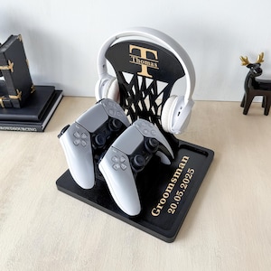 Puede incluir: Un soporte de juego de madera negra con un soporte para auriculares y ranuras para mandos. El soporte contiene unos auriculares blancos y dos mandos grises. El soporte est&aacute; personalizado con el nombre "Thomas" y la palabra "Groomsman" y la fecha "20.05.2025".