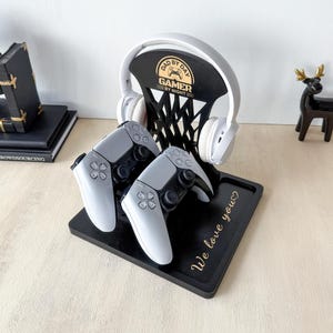 Può includere: Una stazione di gioco in legno nero con un design a canestro da basket, che contiene una cuffia bianca e due controller di gioco grigi. La stazione presenta il testo "DAD BY DAY GAMER BY NIGHT" e "We love you" in lettere dorate.