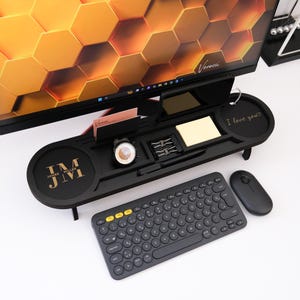 May include: A black desk organizer with a monitor stand, holding a watch, stationery, and a notepad. The organizer has the initials "JM" and the words "James Miller" on it. A wireless keyboard and mouse are in front.