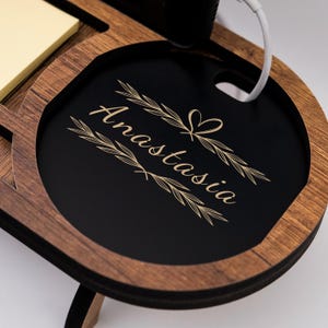 May include: Wooden desk organizer with a black circular tray engraved with the name "Anastasia" in a decorative font. The organizer has a light brown wood finish and includes a section for a notepad and a phone charging port.