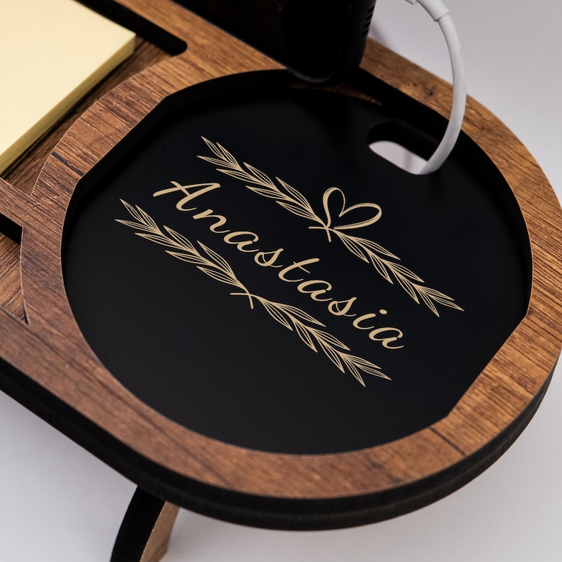 May include: Wooden desk organizer with a black circular tray engraved with the name "Anastasia" in a decorative font. The organizer has a light brown wood finish and includes a section for a notepad and a phone charging port.