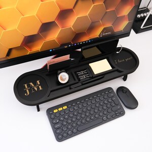 K&ouml;nnte beinhalten: Ein schwarzer Schreibtisch-Organizer mit Monitorst&auml;nder, der eine Uhr, Schreibwaren und einen Notizblock enth&auml;lt. Der Organizer tr&auml;gt die Initialen "JM" und die Worte "James Miller". Eine kabellose Tastatur und Maus befinden sich davor.