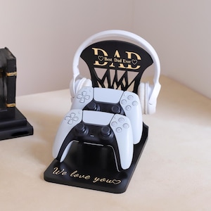 Peut inclure: Support noir pour manettes de jeu avec l'inscription dorée "DAD Best Dad Ever" et "We love you". Le support contient deux manettes blanches et un casque blanc.