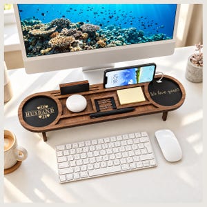 May include: A dark brown wooden desk organiser with a horizontal rectangular shape and rounded ends. It features a smartphone stand, a pen slot, a sticky note compartment, and two circular black insets. One inset reads "HUSBAND" and the other "We love you".