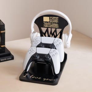 May include: A black wooden gaming stand with the text "I love you" and a design that holds two white game controllers and a pair of white headphones. The stand has the text "Best Dad Ever" and an American flag design.