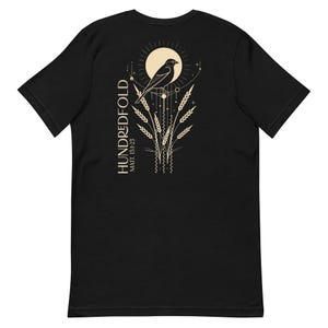 May include: Black t-shirt featuring a gold design of a bird perched on wheat stalks with a sun and stars. The text "HUNDREDFOLD" and "MATT. 13:12-23" is printed on the shirt.