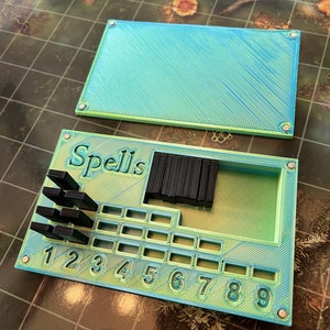 May include: A green and blue plastic game board with the word "Spells" printed on it. The board has a grid of squares with numbers 1 through 9 printed below them. There are black plastic pieces in the top left and right corners of the board.