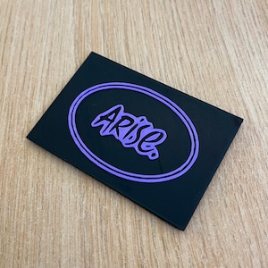 May include: A black rectangular tag with a purple oval border. The word "ARISE" is written in a stylised font inside the oval.