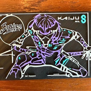 Peut inclure: Une illustration en noir et blanc d'un Kaiju, un monstre fictif, avec des accents violets et bleus. L'illustration est sur un fond noir avec le texte "Kaiju No. 8" en blanc.