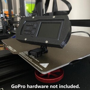 May include: Black control panel with buttons and a screen, mounted on a 3D printer bed. The printer bed is black with a red wheel. The text "GoPro hardware not included" is at the bottom.