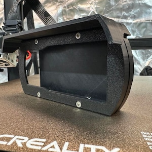 Può includere: Un involucro nero per stampante 3D con una finestra rettangolare e bordi arrotondati. L'involucro è montato su una superficie marrone con la scritta "CREALITY" stampata in bianco. I componenti della stampante sono visibili.