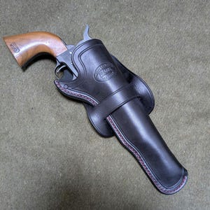 以下が含まれることがあります： 黒い革製のホルスターとそれに合うリボルバー。 ホルスターは縁に赤いステッチがあり、ベルトループが付いています。 リボルバーは木製のグリップと銀色のバレルを持っています。 ホルスターには「FRANK MAKER」と刻印されています。