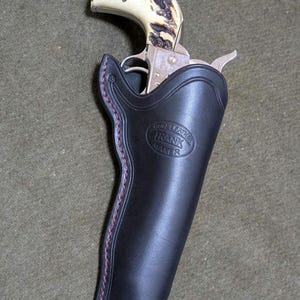 May include: A black leather holster with red stitching, holding a silver revolver with a cream-coloured handle. The holster has the text "GUN LEATHER FRANK MAKER" embossed on it. The revolver is partially visible.