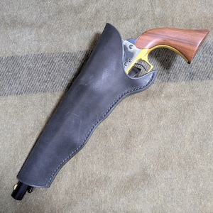 May include: A black leather holster containing a revolver with a polished metal frame, a gold-coloured trigger guard, and a smooth brown wooden grip. The holster features visible stitching along the edges and a small brass-coloured stud at the bottom.