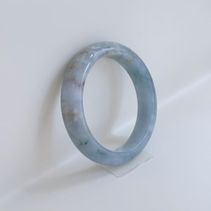Puede incluir: Un brazalete circular liso hecho de piedra pulida. El material presenta una base translúcida de color azul grisáceo pálido con parches blancos moteados y sutiles toques de inclusiones minerales verdes y marrones en toda la superficie.