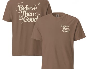 Puede incluir: Dos camisetas marrones. Una muestra la espalda con el texto "Believe There is Good" en una fuente retro color crema, con detalles de estrellas. La otra camiseta muestra el frente con el mismo texto en un tamaño más pequeño.