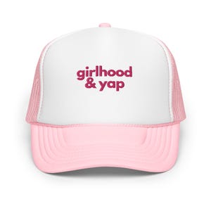 May include: A pink and white trucker hat with the text "girlhood & yap" in a magenta color. The hat has a white front panel, a pink brim, and a pink mesh back.