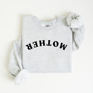 Puede incluir: Una sudadera gris con la palabra "MOTHER" impresa en letras negras en un diseño arqueado.