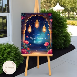 May include: A welcome sign for a Sangeet celebration with a blue and gold archway decorated with hanging lanterns and floral accents. The sign reads "Welcome to Raj & Simran's Sangeet".