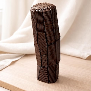 May include: A dark brown, geometric vase constructed from small, rectangular wooden pieces. The vase has a unique, angular design with a textured surface. It stands on a light-colored wooden surface, with a white cloth in the background.