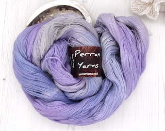 May include: A ball of luxury yarn in shades of lavender, periwinkle, and grey. The yarn is labelled "Perran Yarns" and is displayed on a white wooden surface. The yarn is arranged in a loose, flowing shape, suggesting its softness and texture.