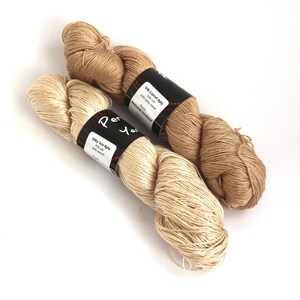 Puede incluir: Dos ovillos de hilo, uno beige y otro crema, con etiquetas que dicen "Silk Camel 4ply 50% Silk 50% Baby Camel Natural Undyed Shade" y "Silky Sea 4ply 70% Silk 30% Sea Silk".