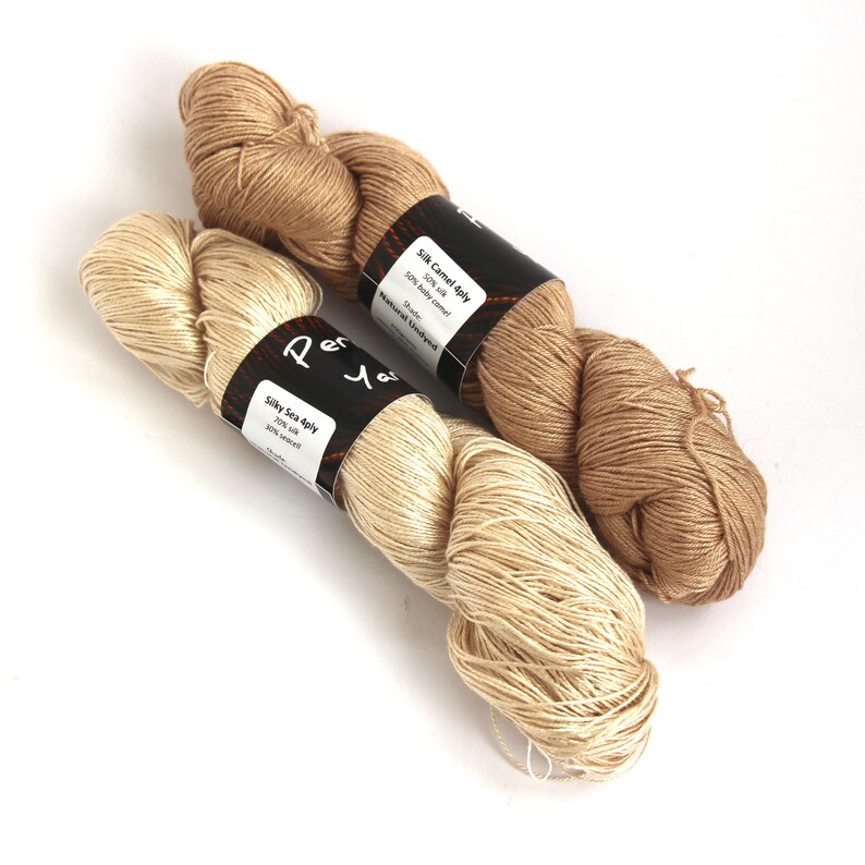 Puede incluir: Dos ovillos de hilo, uno beige y otro crema, con etiquetas que dicen "Silk Camel 4ply 50% Silk 50% Baby Camel Natural Undyed Shade" y "Silky Sea 4ply 70% Silk 30% Sea Silk".