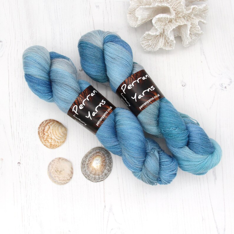 May include: Two skeins of blue variegated yarn with brown labels that read "Perran Yarns Luxury perranyarns.co.uk". The yarn is next to seashells and a white coral decoration. The yarn is a mix of light and dark blues.